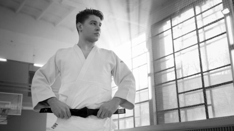 A young martial artist adjusts his black belt in a sunlit gym.