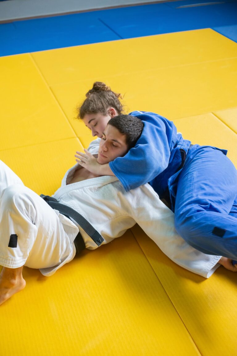 Two adults practicing judo in a grappling hold on a yellow mat indoors.